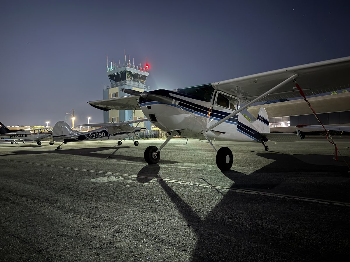A blue-and-white Cessna 170 tied down on the ramp at night, with the lit Jack Northrop Field control tower and other parked aircraft behind it
